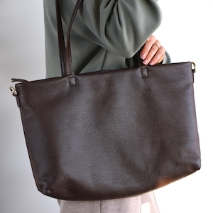 May include: A dark brown leather tote bag with long shoulder straps. The bag has a textured surface and a rectangular shape. The straps are also brown and appear to be made of the same material. The bag is being held by a person.