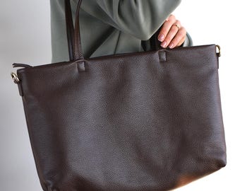 May include: A dark brown leather tote bag with long shoulder straps. The bag has a textured surface and a rectangular shape. The straps are also brown and appear to be made of the same material. The bag is being held by a person.