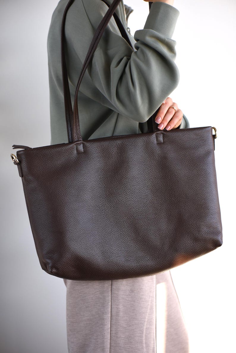 May include: A dark brown leather tote bag with long shoulder straps. The bag has a textured surface and a rectangular shape. The straps are also brown and appear to be made of the same material. The bag is being held by a person.