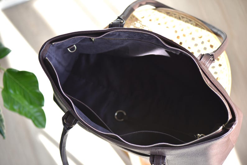 May include: A dark brown leather tote bag with a silver zipper and handles. The interior is black with silver metal rings. The bag is open, revealing the inside. The bag is sitting on a light-colored surface.