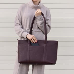 May include: A deep burgundy tote bag with a rectangular shape and two long handles. The bag has a pocket on the front and is held by a person wearing sunglasses and a matching outfit.