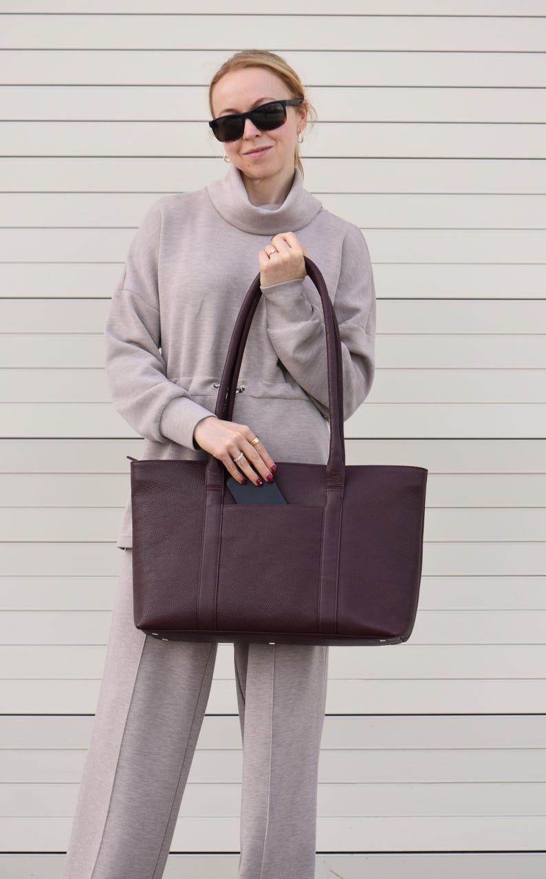 May include: A deep burgundy tote bag with a rectangular shape and two long handles. The bag has a pocket on the front and is held by a person wearing sunglasses and a matching outfit.