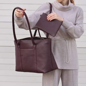 May include: A burgundy leather tote bag with long handles and a matching small pouch. The tote has a rectangular shape with a zippered top and an exterior pocket. The pouch has a zipper closure. The bags are a deep, rich color.