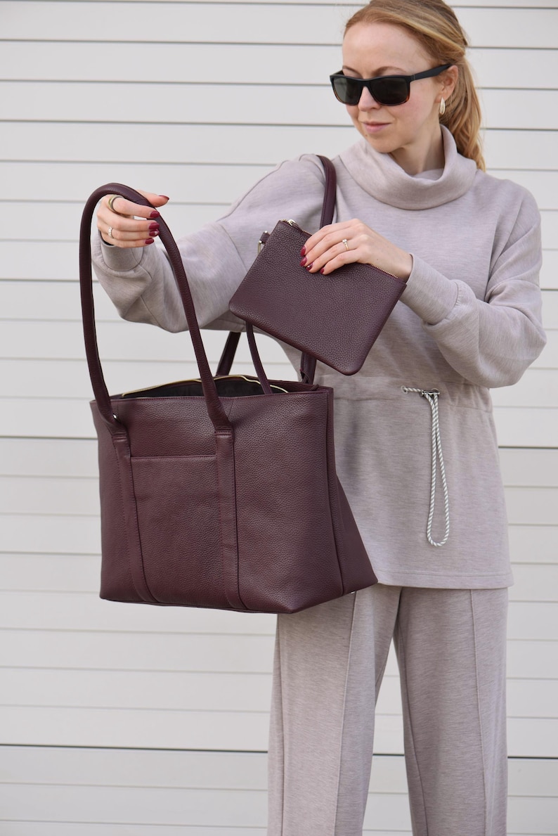 May include: A burgundy leather tote bag with long handles and a matching small pouch. The tote has a rectangular shape with a zippered top and an exterior pocket. The pouch has a zipper closure. The bags are a deep, rich color.