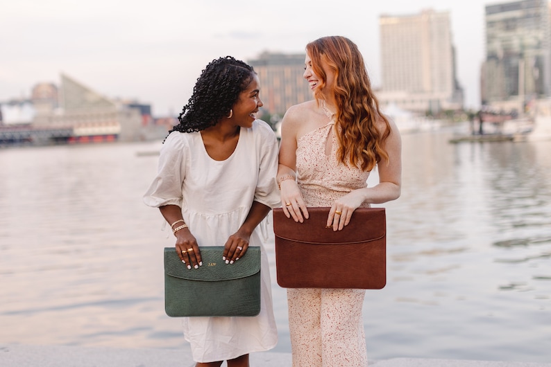 Lifestyle photo of two women holding leather laptop sleeves— Aloe green and Coffee brown—personalized monograms, professional work accessories