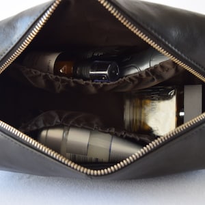 May include: Black leather toiletry bag with a gold zipper, open to reveal various grooming products. The bag has a handle and a brown interior with pockets. The contents include bottles, jars, and a metal container.