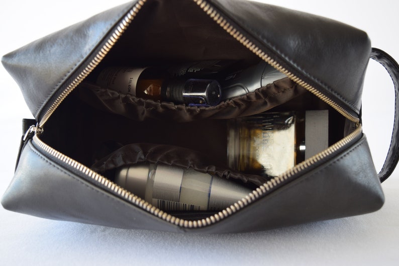 May include: Black leather toiletry bag with a gold zipper, open to reveal various grooming products. The bag has a handle and a brown interior with pockets. The contents include bottles, jars, and a metal container.