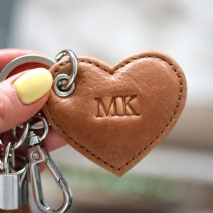 May include: A heart-shaped tan leather keychain featuring the embossed letters MK in the center. The leather heart is attached to a silver-toned metal split ring and a lobster claw clasp by a small metal grommet and ring hardware.