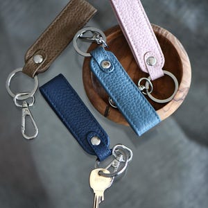 Op de afbeelding: Vier leren sleutelhangers in bruin, blauw en roze. Elke sleutelhanger heeft een zilveren metalen ring en een drukknoopsluiting. E&eacute;n sleutelhanger heeft een zilveren karabijnhaak. De sleutelhangers liggen op een reflecterend oppervlak.