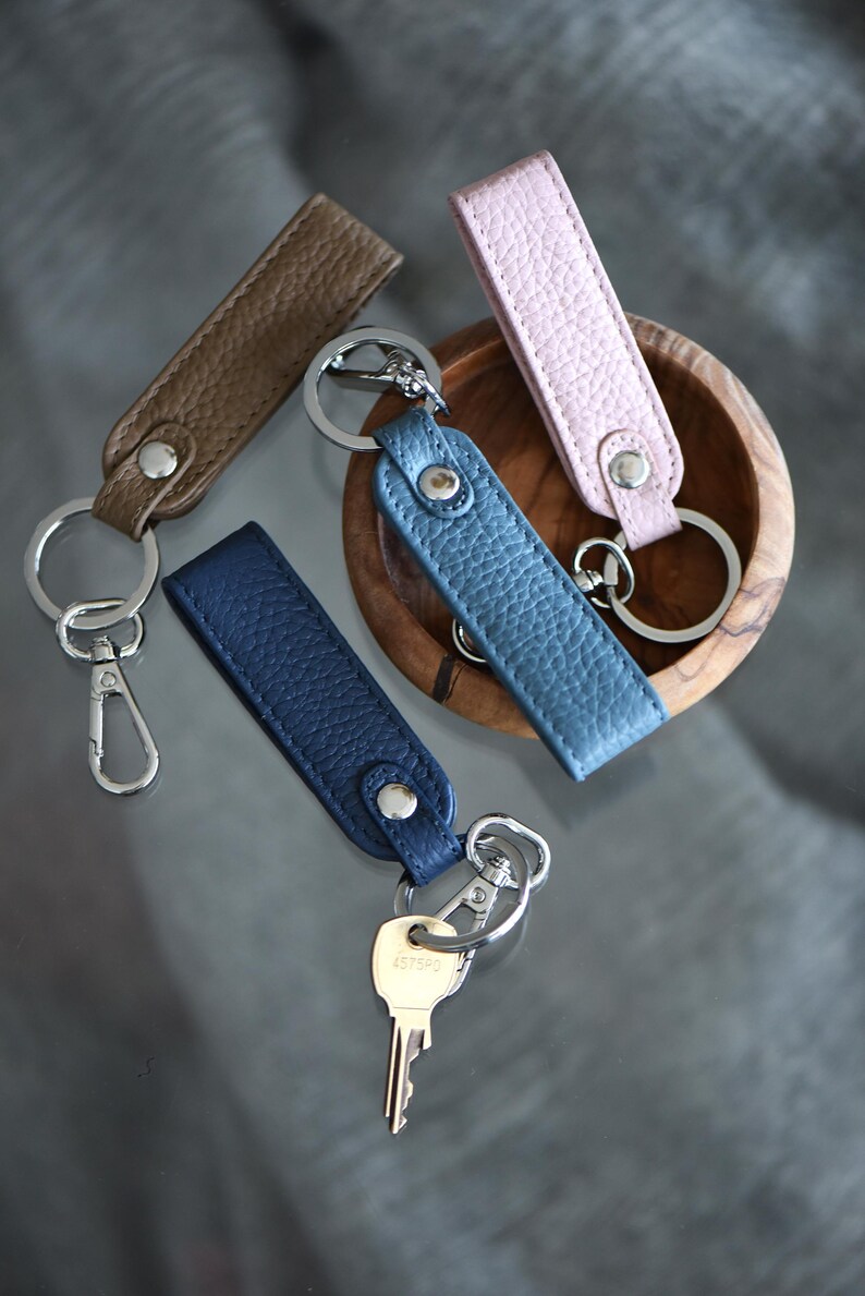 Op de afbeelding: Vier leren sleutelhangers in bruin, blauw en roze. Elke sleutelhanger heeft een zilveren metalen ring en een drukknoopsluiting. E&eacute;n sleutelhanger heeft een zilveren karabijnhaak. De sleutelhangers liggen op een reflecterend oppervlak.
