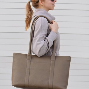 May include: A taupe-colored tote bag with long handles. The bag is rectangular with a textured surface. The person is wearing a gray sweatshirt and sunglasses.