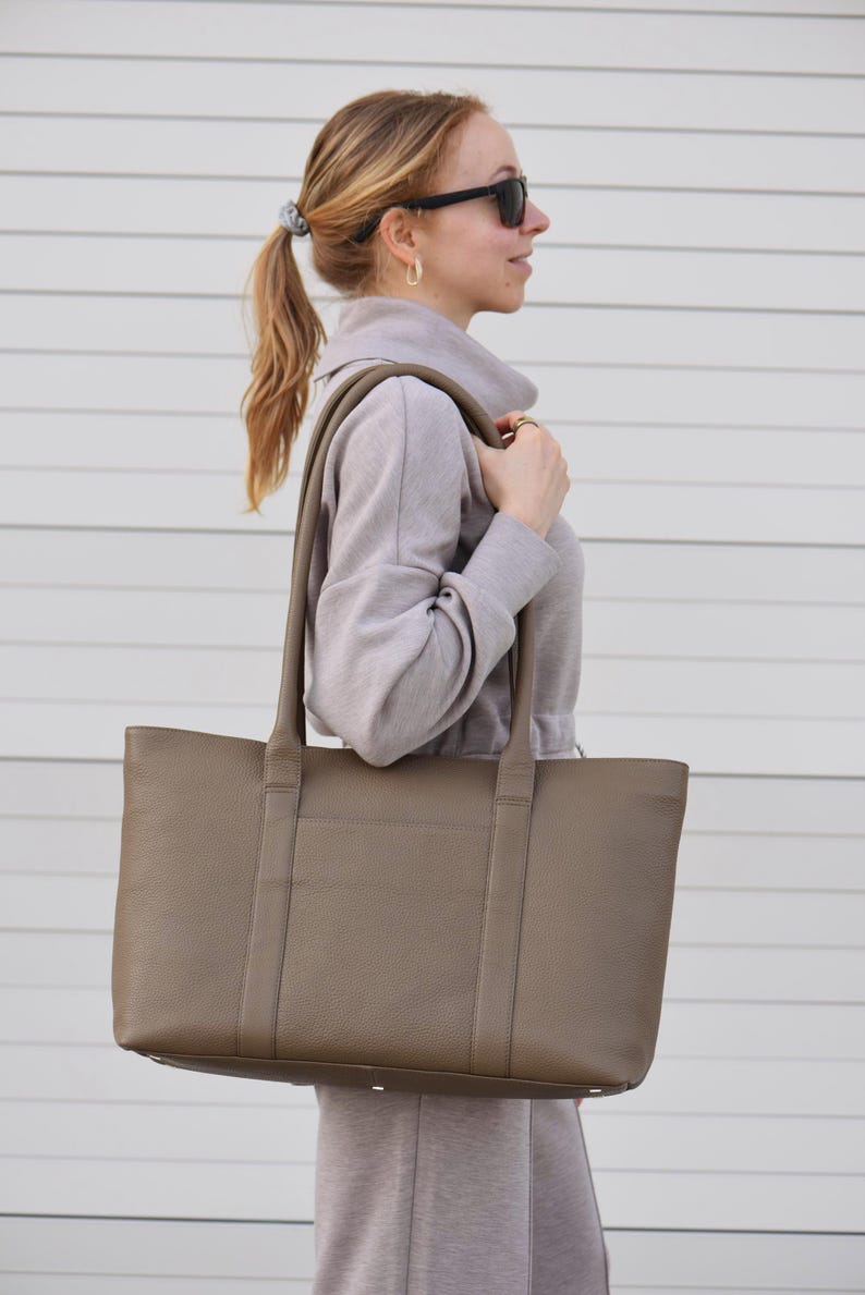 May include: A taupe-colored tote bag with long handles. The bag is rectangular with a textured surface. The person is wearing a gray sweatshirt and sunglasses.