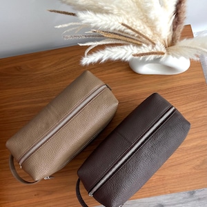 May include: Two leather travel cases in tan and dark brown, each with a silver zipper and a small handle. The cases are rectangular and sit on a wooden surface. A vase of dried flowers is in the background.