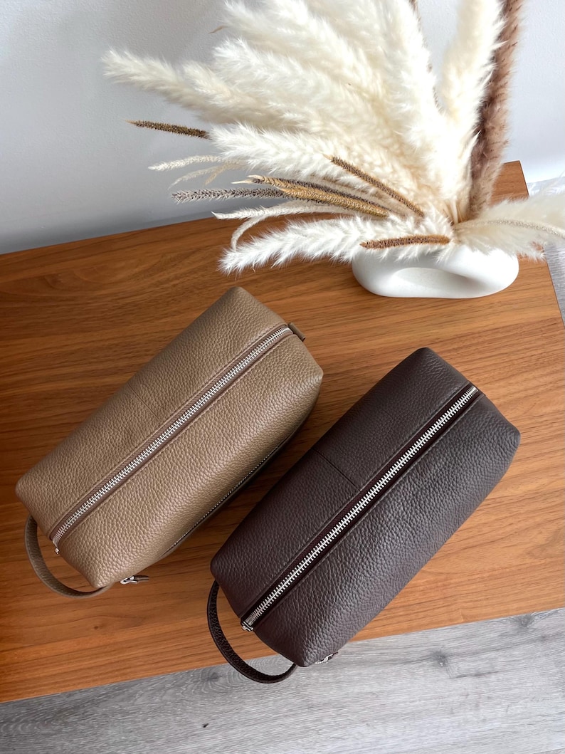 May include: Two leather travel cases in tan and dark brown, each with a silver zipper and a small handle. The cases are rectangular and sit on a wooden surface. A vase of dried flowers is in the background.