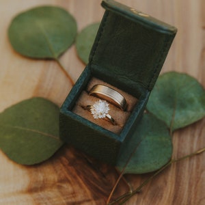 May include: Two gold wedding bands in a green velvet ring box. The box is open and the rings are visible. The box is sitting on a wooden surface with green leaves.