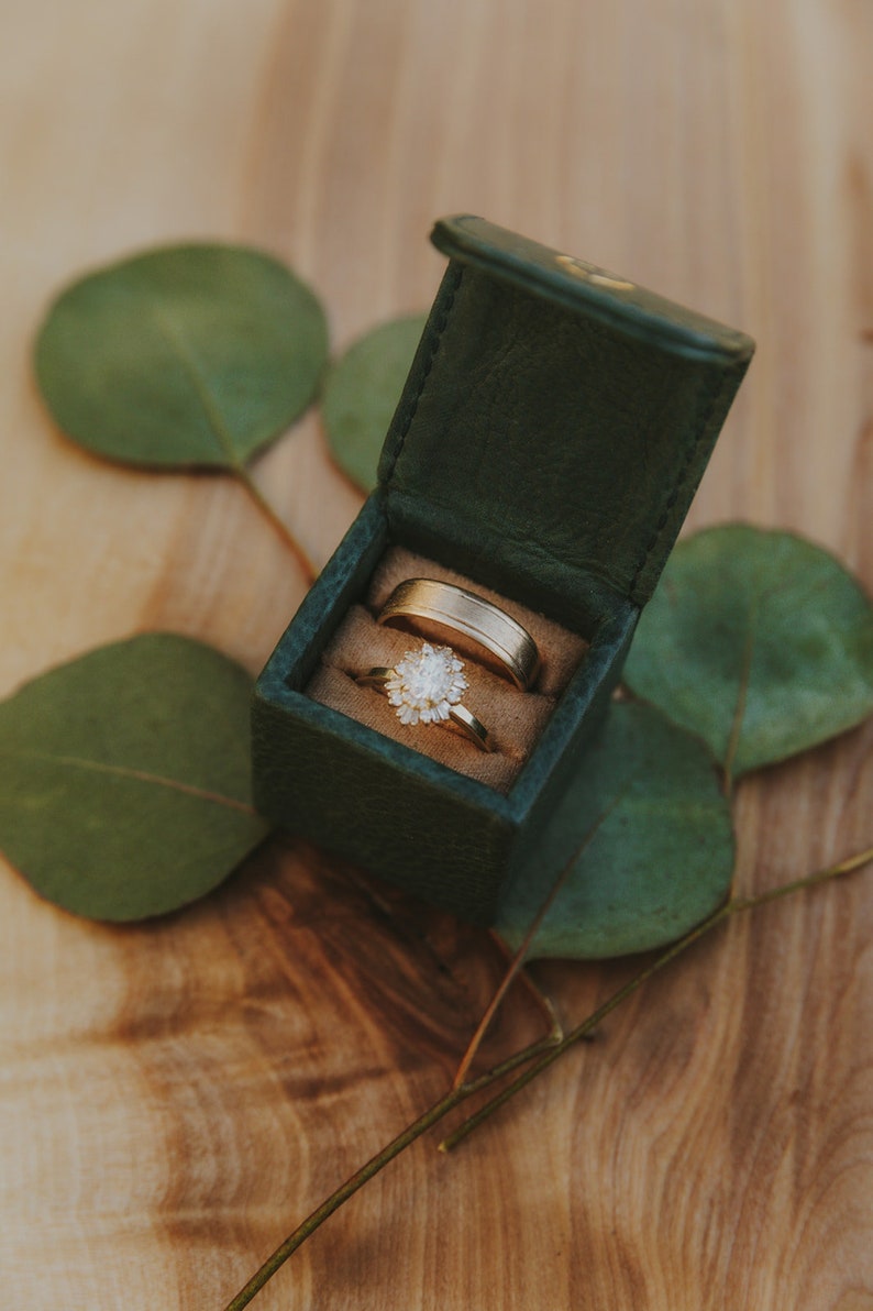 May include: Two gold wedding bands in a green velvet ring box. The box is open and the rings are visible. The box is sitting on a wooden surface with green leaves.