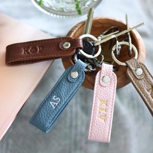 Op de afbeelding: Gepersonaliseerde leren sleutelhangers in bruin, blauw en roze. Elke sleutelhanger heeft initialen en is bevestigd aan een metalen ring. De sleutelhangers worden in een houten kom getoond. De sleutelhangers zijn ongeveer 10 cm lang.