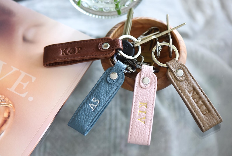 Op de afbeelding: Gepersonaliseerde leren sleutelhangers in bruin, blauw en roze. Elke sleutelhanger heeft initialen en is bevestigd aan een metalen ring. De sleutelhangers worden in een houten kom getoond. De sleutelhangers zijn ongeveer 10 cm lang.