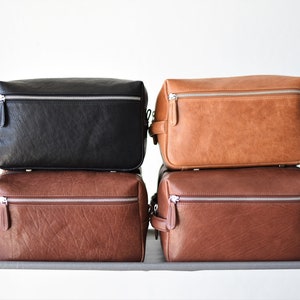 May include: Four rectangular toiletry bags in black, tan, and two shades of brown are stacked. Each bag has a silver zipper closure and a small handle on one side. The bags are made of a textured material.