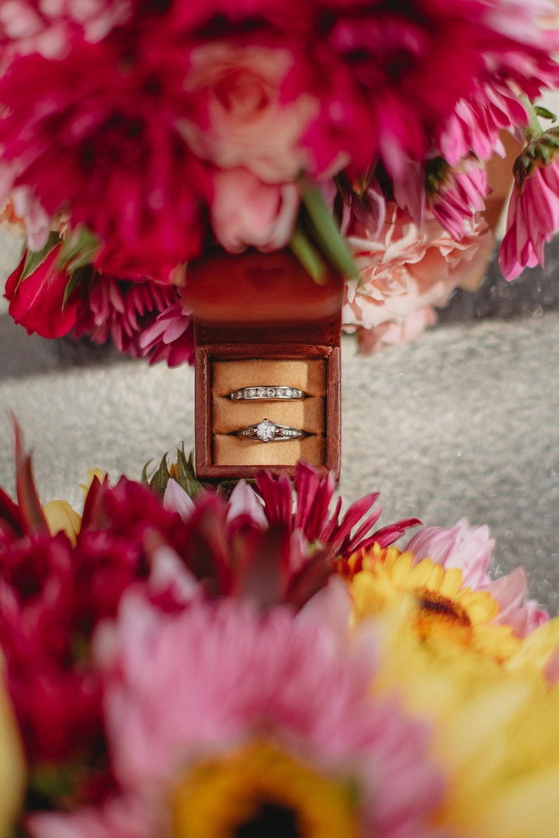 May include: Two wedding bands in a small brown velvet ring box surrounded by pink and yellow flowers.