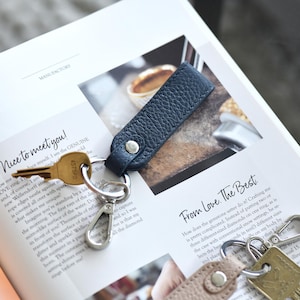Op de afbeelding: Een marineblauwe leren sleutelhanger met een zilveren karabijnhaak en een gouden sleutel. De sleutelhanger is bevestigd aan een sleutel. De tekst "Nice to meet you!" is zichtbaar op het open boek.
