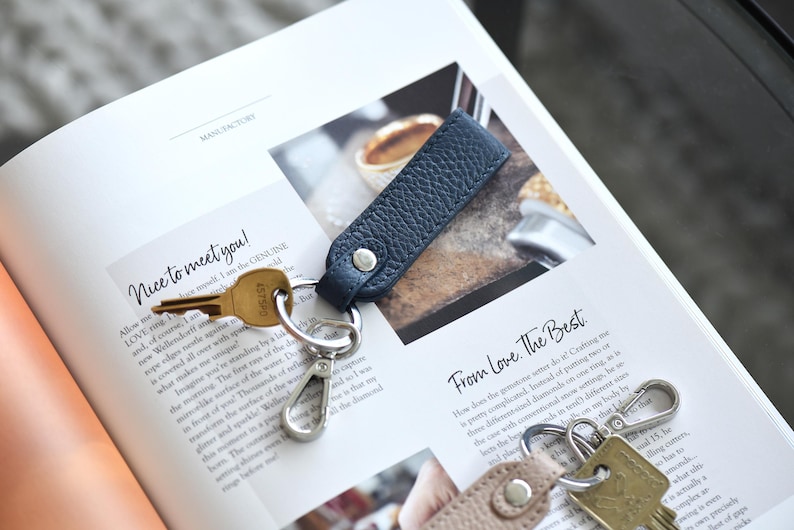 Op de afbeelding: Een marineblauwe leren sleutelhanger met een zilveren karabijnhaak en een gouden sleutel. De sleutelhanger is bevestigd aan een sleutel. De tekst "Nice to meet you!" is zichtbaar op het open boek.