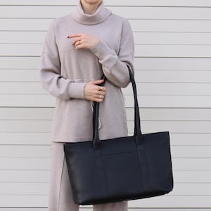 May include: A black tote bag with long handles. The bag is rectangular and appears to be made of leather. The person is wearing a light gray turtleneck sweater and matching pants.
