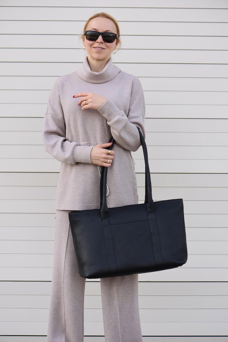 May include: A black tote bag with long handles. The bag is rectangular and appears to be made of leather. The person is wearing a light gray turtleneck sweater and matching pants.