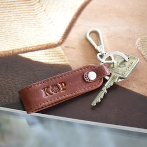 Op de afbeelding: Een bruin leren sleutelhanger met de initialen "KOP" erin ge&euml;mbosseerd. Het heeft een zilveren sluiting en een sleutel. De achtergrond is een tijdschrift met een bruin en beige patroon.