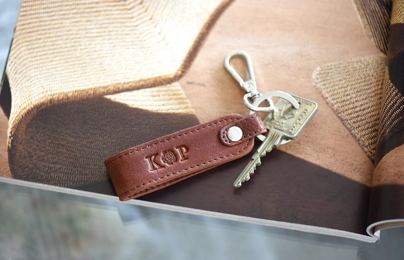Op de afbeelding: Een bruin leren sleutelhanger met de initialen "KOP" erin ge&euml;mbosseerd. Het heeft een zilveren sluiting en een sleutel. De achtergrond is een tijdschrift met een bruin en beige patroon.