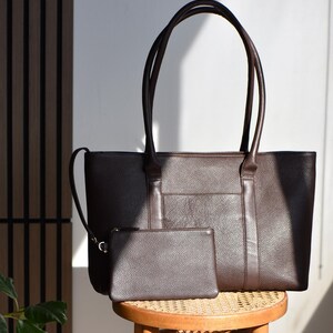 May include: A brown leather tote bag with matching small pouch. The tote has long handles and a front pocket. The pouch has a zipper closure. Both items are sitting on a small wooden stool with a woven seat.