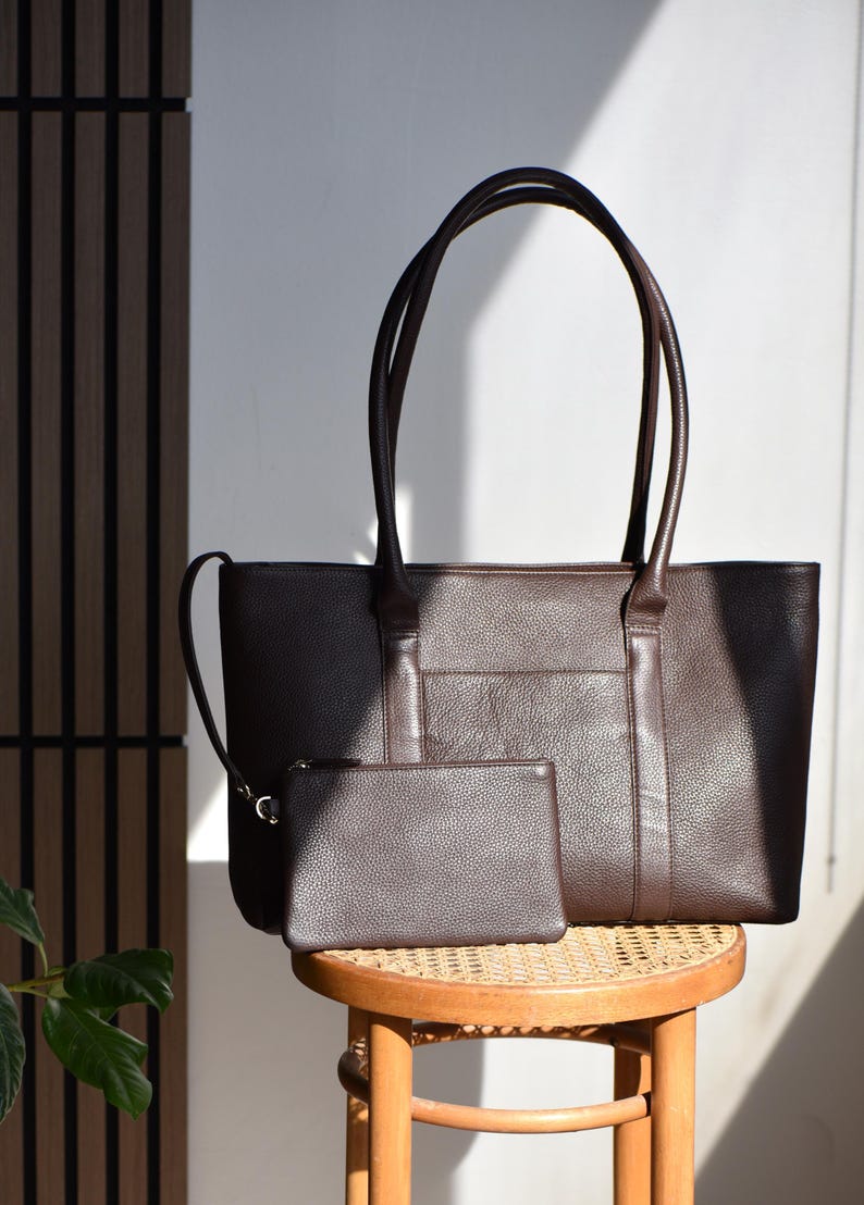 May include: A brown leather tote bag with matching small pouch. The tote has long handles and a front pocket. The pouch has a zipper closure. Both items are sitting on a small wooden stool with a woven seat.