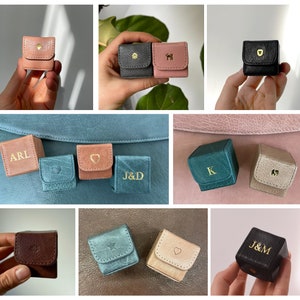May include: An assortment of small, personalised leather boxes in various colours, including brown, black, pink, blue, and green. Each box features a different letter or symbol, such as "ARL", "J&D", "K", and a heart.