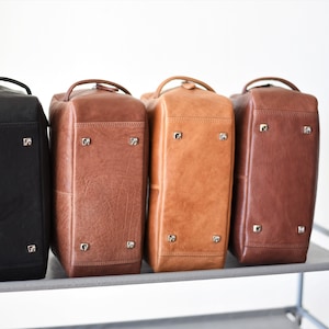 May include: Four leather travel bags in black, brown, tan, and dark brown are displayed side-by-side. Each bag has a handle, metal feet, and a rectangular shape. The bags are on a gray shelf against a white background.