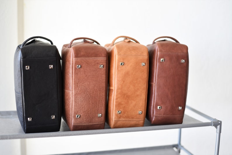 May include: Four leather travel bags in black, brown, tan, and dark brown are displayed side-by-side. Each bag has a handle, metal feet, and a rectangular shape. The bags are on a gray shelf against a white background.