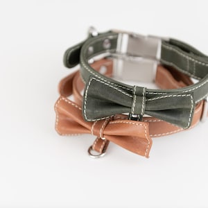 May include: Two leather dog collars with bow ties. The top collar is green with white stitching and a silver buckle. The bottom collar is brown with white stitching and a silver D-ring.