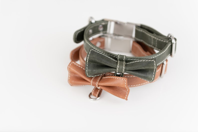 May include: Two leather dog collars with bow ties. The top collar is green with white stitching and a silver buckle. The bottom collar is brown with white stitching and a silver D-ring.