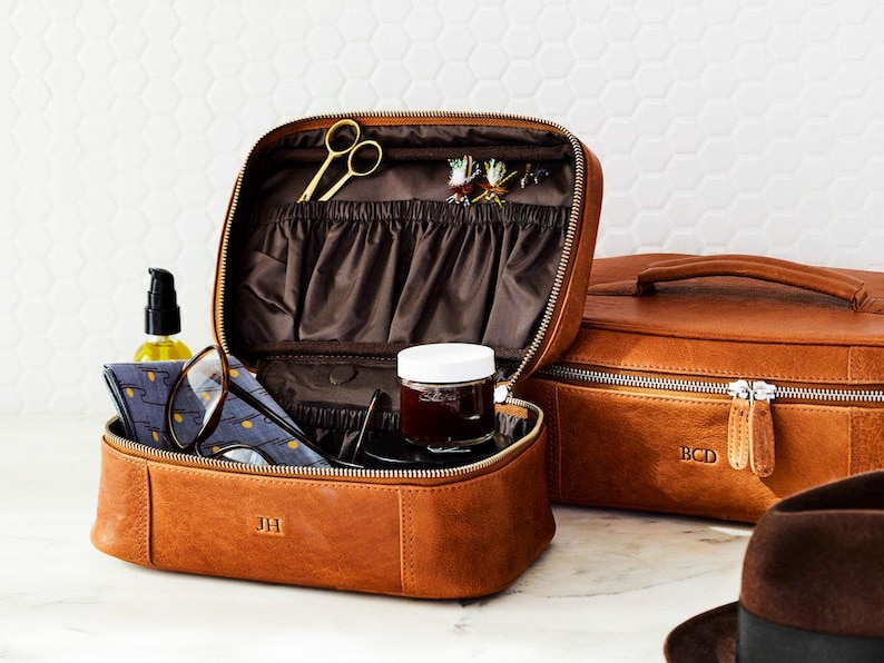 May include: A tan leather travel case with a zipper closure, featuring the initials JH embossed on the front. The interior is lined in dark brown fabric with elastic pockets, gold scissors, and a small jar. A second matching case shows BCD.