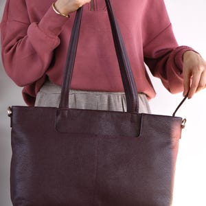 May include: A large, burgundy leather tote bag with long handles. The bag has a rectangular shape and a textured surface. The handles are attached with gold-colored hardware. The bag is held by a person wearing a mauve sweatshirt and gray pants.