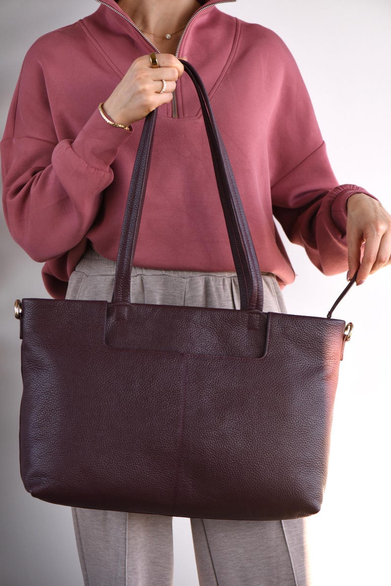 May include: A large, burgundy leather tote bag with long handles. The bag has a rectangular shape and a textured surface. The handles are attached with gold-colored hardware. The bag is held by a person wearing a mauve sweatshirt and gray pants.