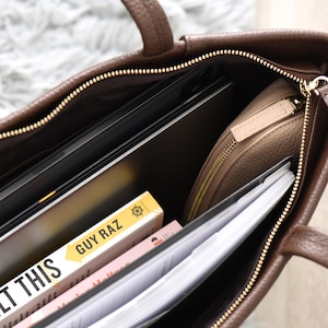 May include: A brown leather tote bag with a gold zipper, handles, and hardware. The bag is open, revealing a laptop, a beige zippered pouch, books, and papers. The book title "I BUILT THIS" is visible.