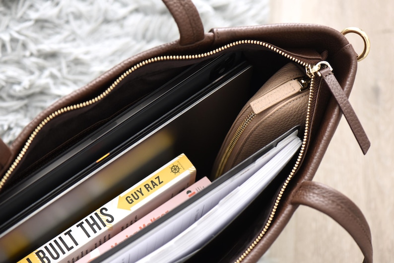 May include: A brown leather tote bag with a gold zipper, handles, and hardware. The bag is open, revealing a laptop, a beige zippered pouch, books, and papers. The book title "I BUILT THIS" is visible.