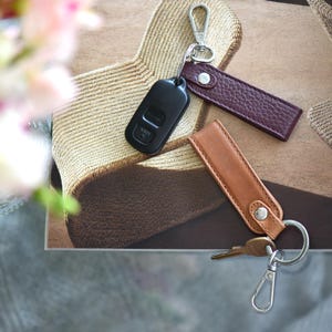 Op de afbeelding: Een bovenaanzicht van sleutelhangers en een autosleutel. De sleutelhangers zijn gemaakt van bruin en bordeauxrood leer, met zilverkleurige sluitingen. Er is ook een zwarte autosleutel aanwezig. De items zijn gerangschikt op een patroon.