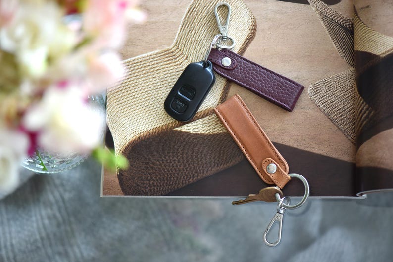 Op de afbeelding: Een bovenaanzicht van sleutelhangers en een autosleutel. De sleutelhangers zijn gemaakt van bruin en bordeauxrood leer, met zilverkleurige sluitingen. Er is ook een zwarte autosleutel aanwezig. De items zijn gerangschikt op een patroon.