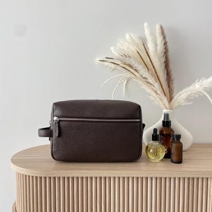 May include: A dark brown leather toiletry bag with a silver zipper and handle sits on a light wood table. Beside it are several small bottles and a white vase with decorative dried plants. The table has a ribbed design.