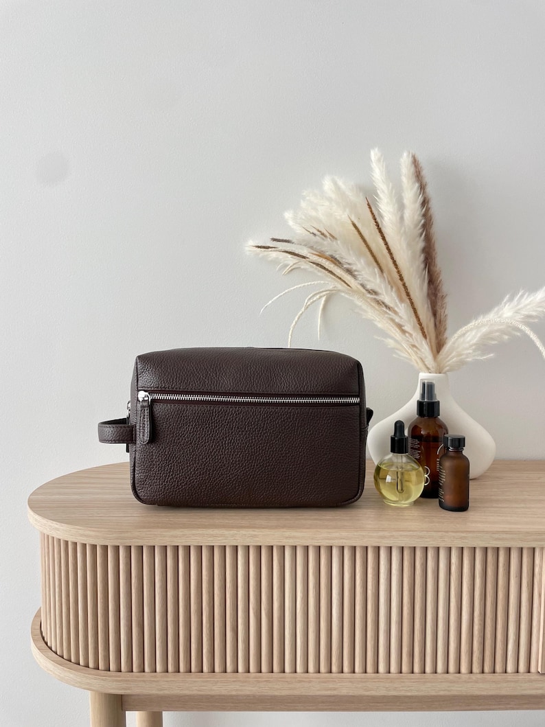 May include: A dark brown leather toiletry bag with a silver zipper and handle sits on a light wood table. Beside it are several small bottles and a white vase with decorative dried plants. The table has a ribbed design.