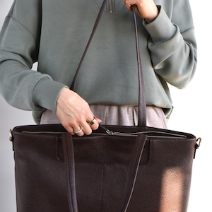 May include: A dark brown leather tote bag with long handles and a zipper closure. The bag has a textured surface and a rectangular shape. The bag is held by a person wearing a green sweatshirt.