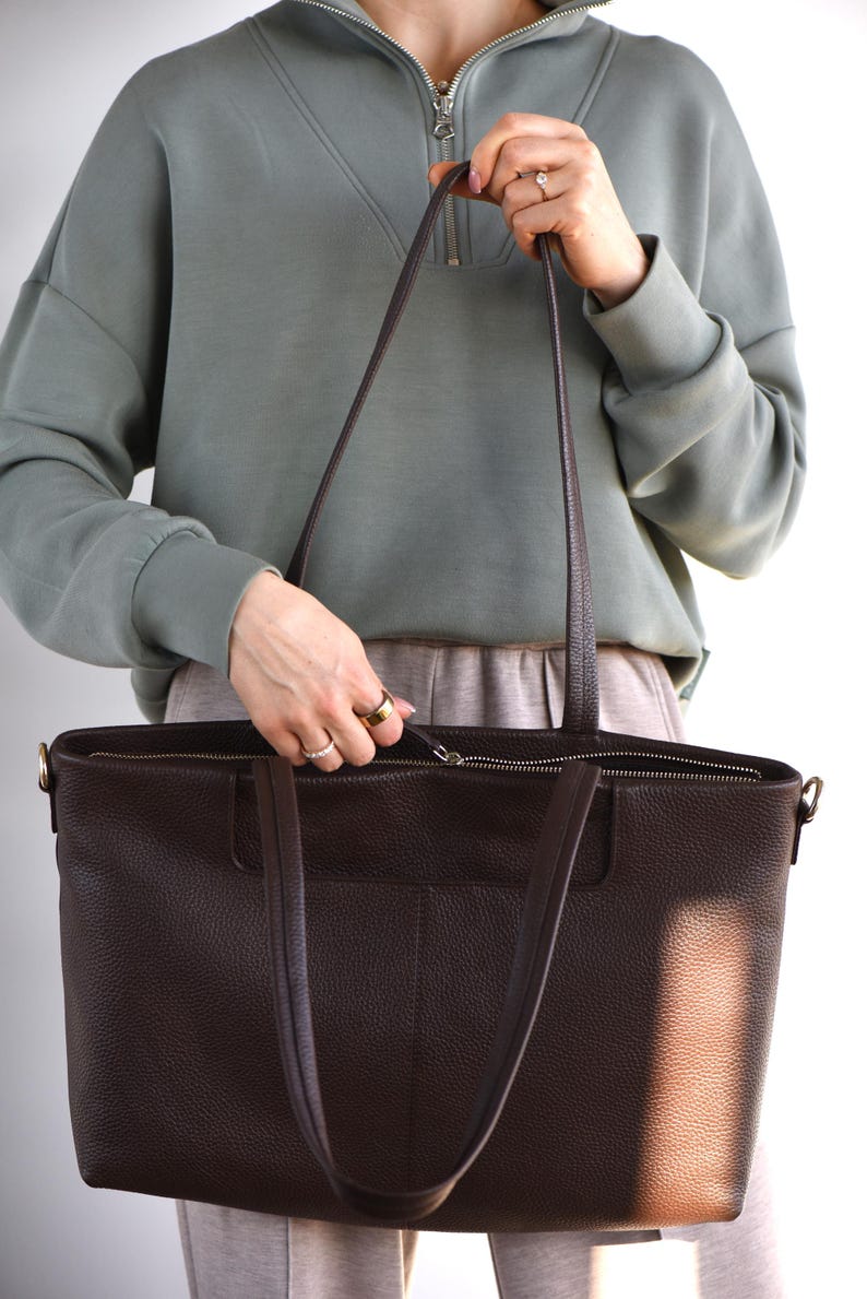 May include: A dark brown leather tote bag with long handles and a zipper closure. The bag has a textured surface and a rectangular shape. The bag is held by a person wearing a green sweatshirt.