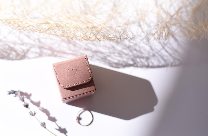 May include: A small, pale pink, square-shaped ring box with a heart embossed on the top. The box is sitting on a white surface with a ring and a sprig of lavender.