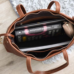 May include: A brown leather tote bag with a zipper closure, containing a laptop, a book titled "How Infrastructure Works," and a small beige wallet. A set of keys with a car remote is visible next to the bag.